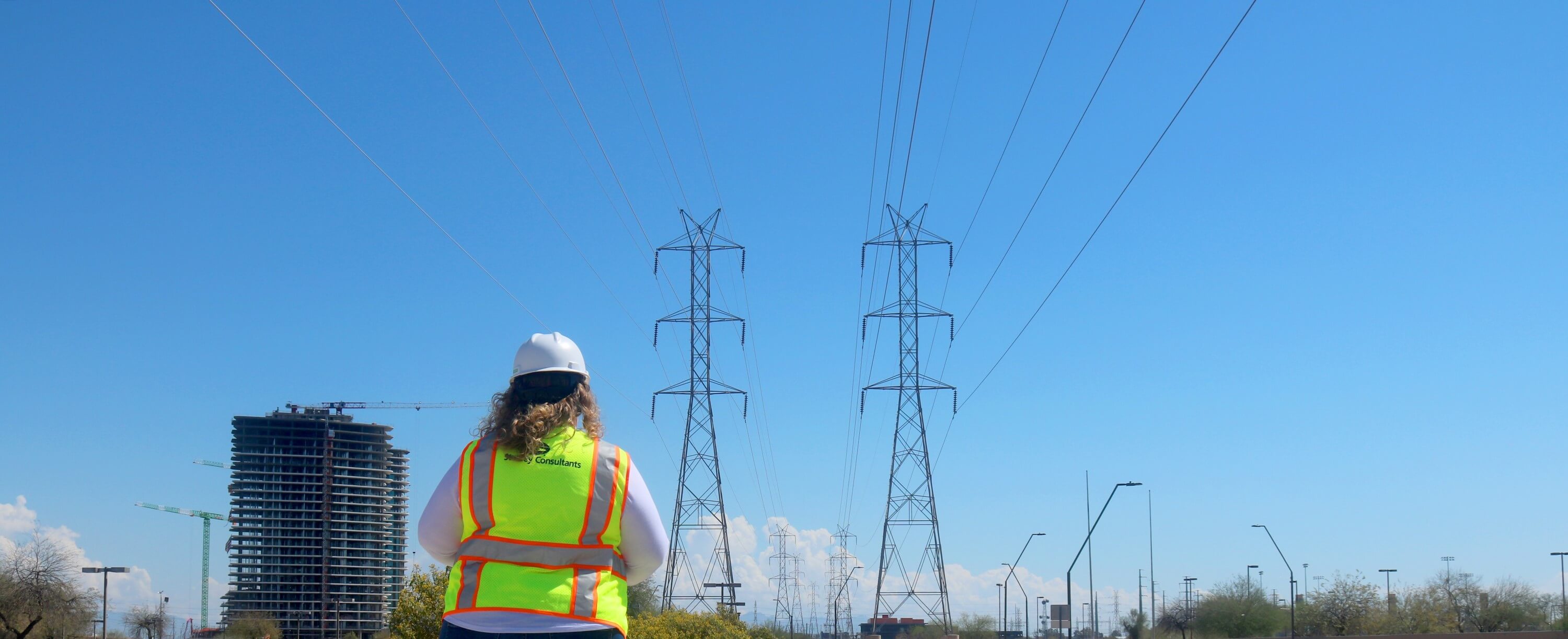 A female engineer wearing a high-visibility vest and helmet, standing in front of electrical transmission towers at an energy infrastructure site.